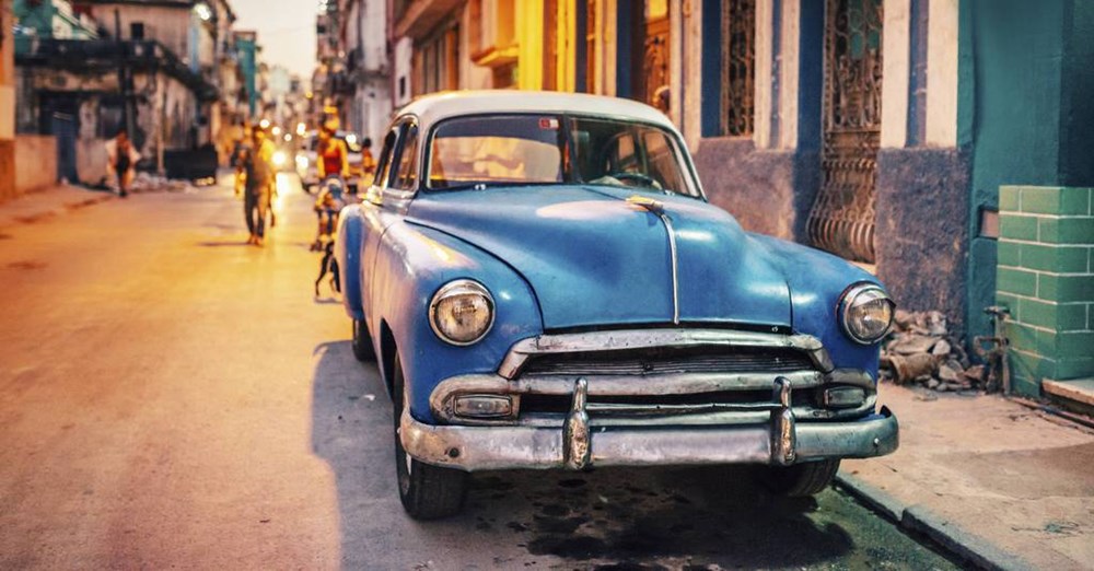 En blå, klassisk bil parkeret på en livlig gade i skumringen med mennesker i baggrunden.