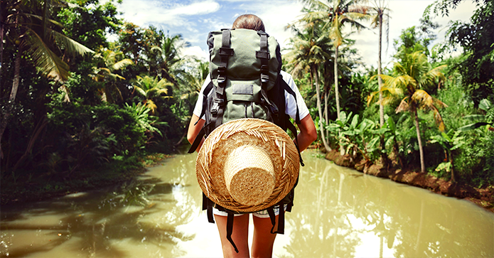 En rygsækrejsende med en stor rygsæk og stråhat står ved en flod omgivet af frodig, tropisk vegetation.