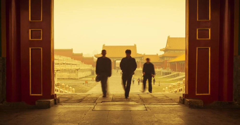 Tre personer går gennem en stor åben port; et glimt af traditionelle kinesiske bygninger ses i baggrunden, og tågen skaber en stemningsfuld atmosfære.