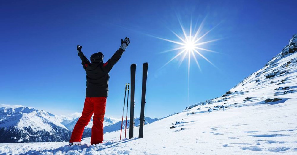 En person i skitøj står med hævede arme på et sneklædt bjerg og nyder solskinnet. Vedkommendes ski står vertikalt i sneen.
