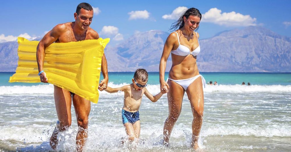En familie nyder en dag på stranden; en mand og en kvinde løber med et grin på grunt vand med en lille dreng, mens manden holder en gul luftmadras.