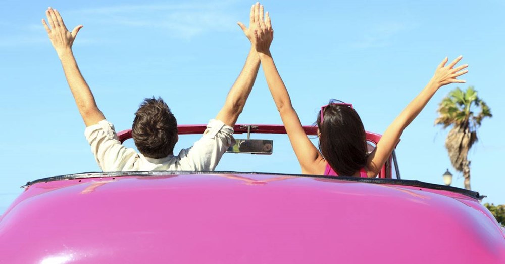 To personer jubler med armene i vejret fra bagagerummet af en lyserød cabriolet, foran palmer og en klar blå himmel.
