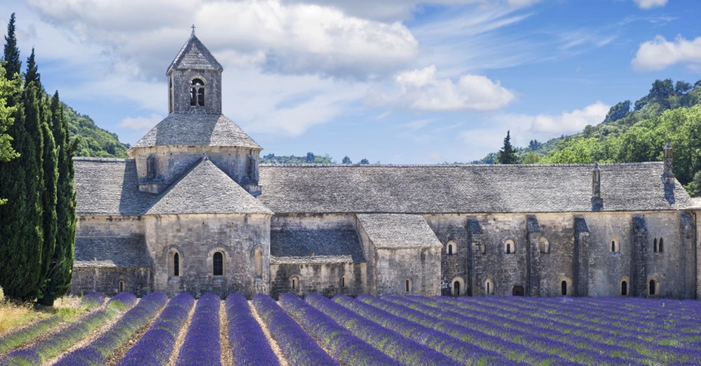 Billede af et historisk stenbygget kloster med en mark af lilla lavendel under en blå himmel.