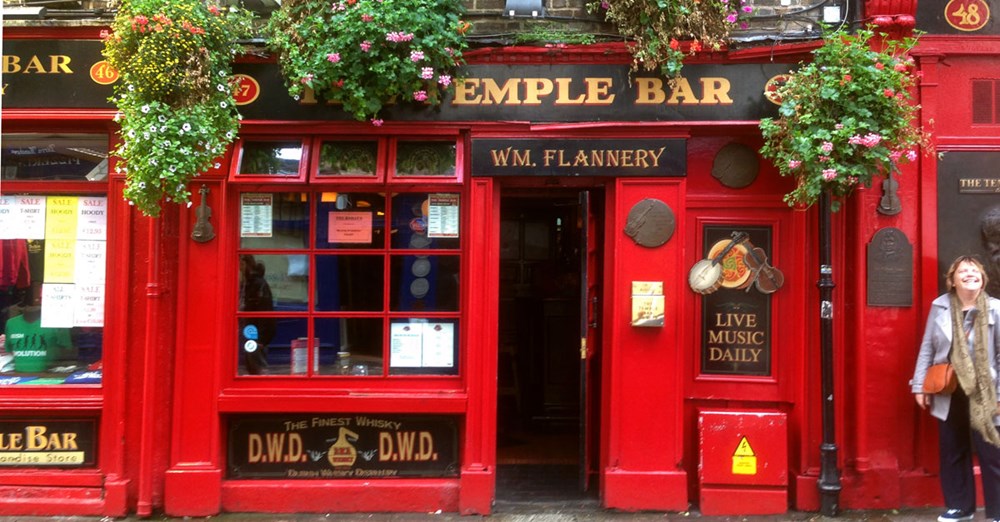 Den ikoniske røde facade af Temple Bar pubben i Dublin, med blomsterkasser og en smilende kvinde foran.