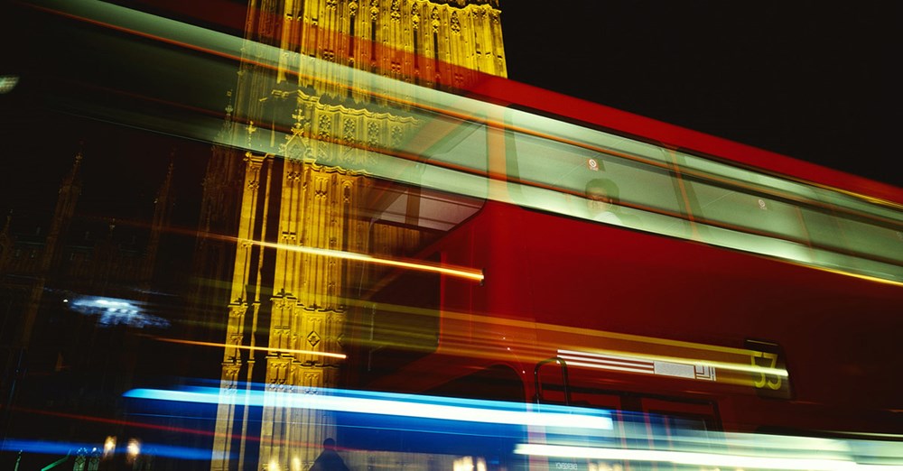 Billedet viser en dynamisk scene med lysstrøg fra en rød bus i bevægelse foran et oplyst historisk bygningsværk om natten.