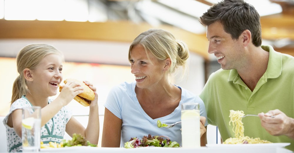 En familie nyder et måltid sammen; en pige spiser en burger, mens moderen og faderen smiler og har salat og pasta på deres tallerkener.