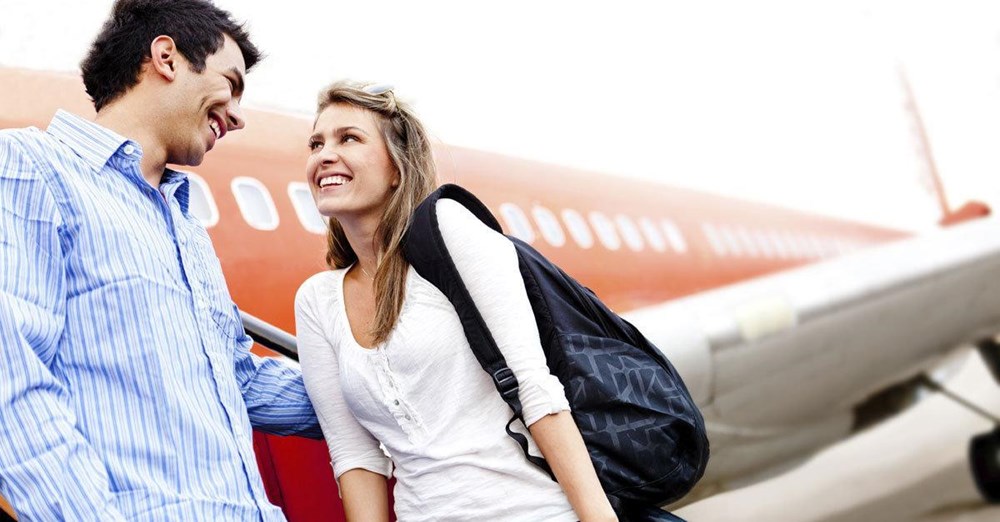 En mand og en kvinde smiler til hinanden med en parkeret flyvemaskine i baggrunden. De bærer afslappet tøj og ser ud til at være i godt humør.