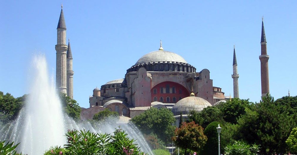 Hagia Sophia i Istanbul, en historisk bygning med kupler og minareter, set gennem vand fra en springvand og omgivet af grønne træer.