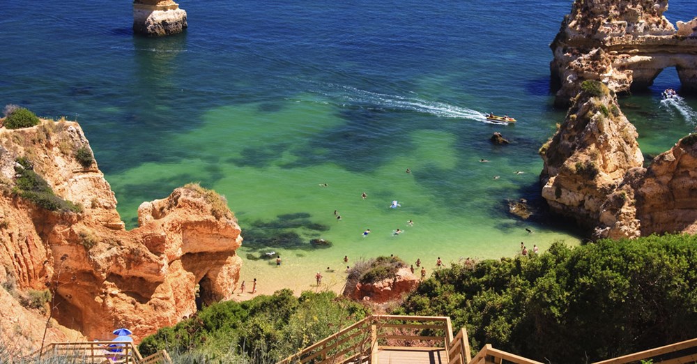 En billedskøn strand med klart blågrønt vand omgivet af klipper. Mennesker bader og soler sig, mens andre går ned af en trætrappe til stranden.