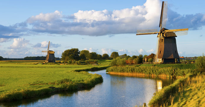 Idyllisk hollandsk landskab med traditionelle vindmøller ved en flod omgivet af grønne enge under en skyet himmel.