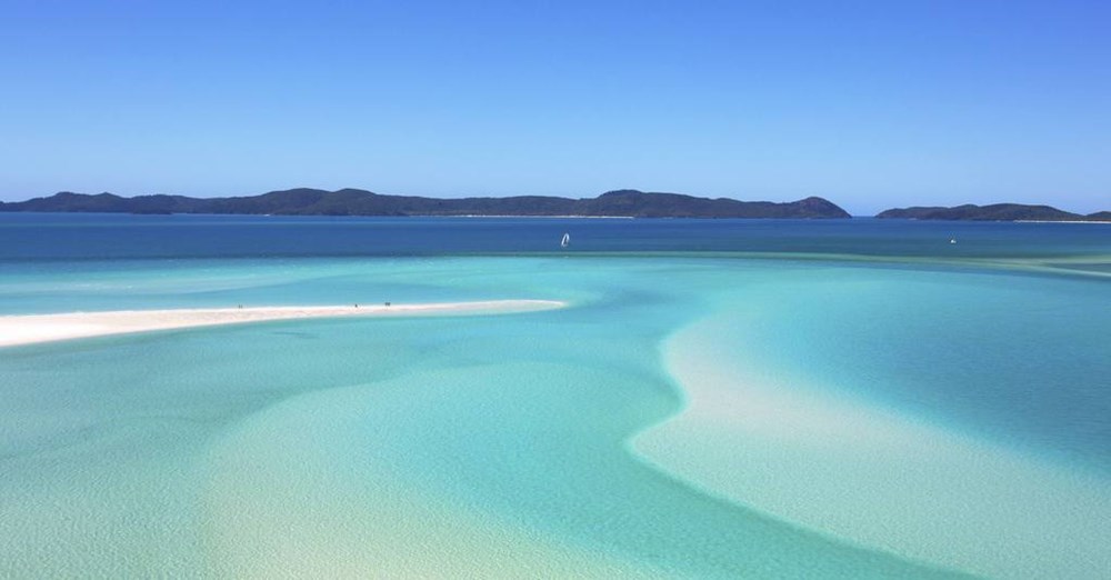 En idyllisk strand med krystalklart, turkisblåt vand og en hvid sandbanke, der bugter sig ind i det rolige hav mod en baggrund af bakkede øer.