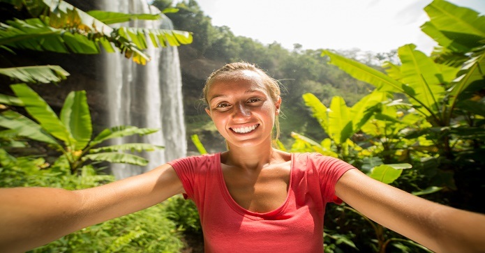 En glad ung kvinde tager et selfie foran et vandfald på Costa Rica omgivet af grønne planter under solskin.