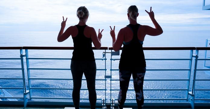 To kvinder står med ryggen til kameraet og laver sejrstegn, mens de kigger ud over havet fra et skibs dæk.
