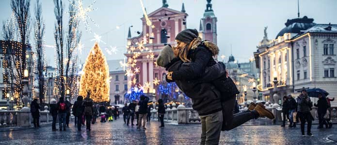 Par kysser hinanden på gade med stort juletræ og smukke bygninger