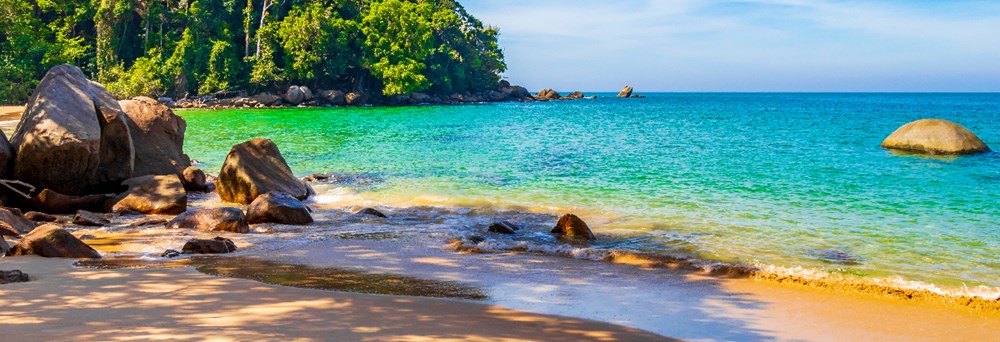 En tropisk strand med klart blågrønt vand, gyldent sand, og klipper ved en frodig grøn skovkant.